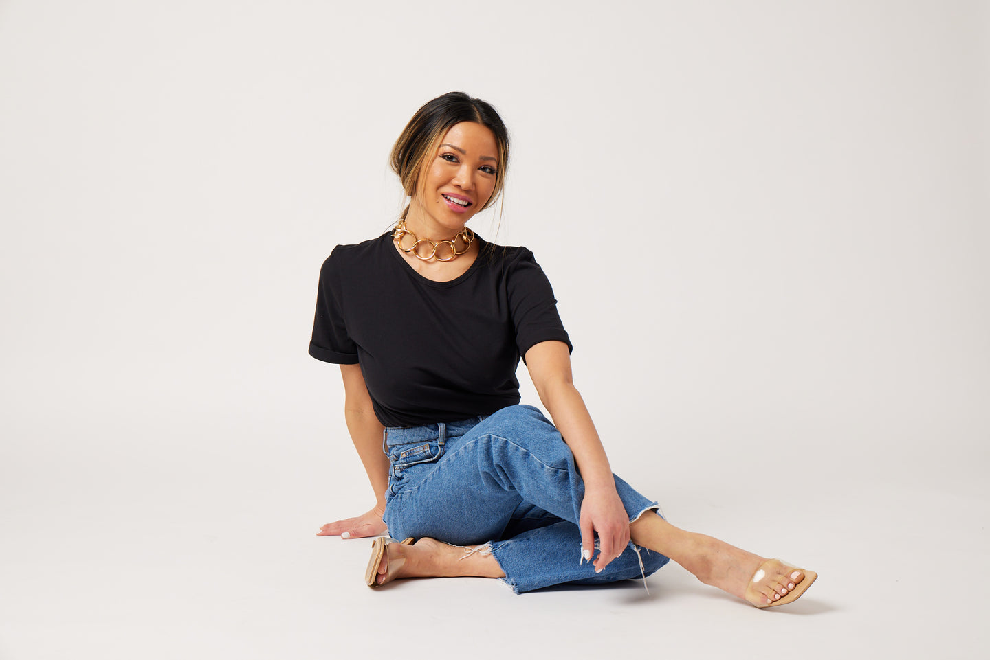 Asian woman wearing a black t-shirt, jeans and a statement necklace in clear sandals sitting on the floor