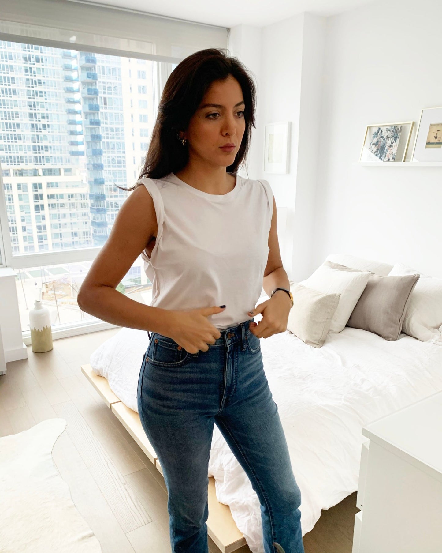 Thin, white model with brunette hair wearing a white pima cotton muscle tee and blue jeans posing in city apartment bedroom