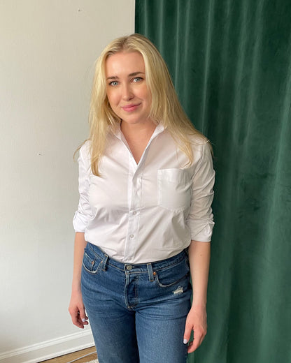 White organic cotton long sleeve button down on blonde white model tucked into blue jeans in front of green curtains