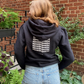 Back detail of black cotton fleece puff sleeve sweatshirt on model wearing blue jean shorts in front of brick wall and plants