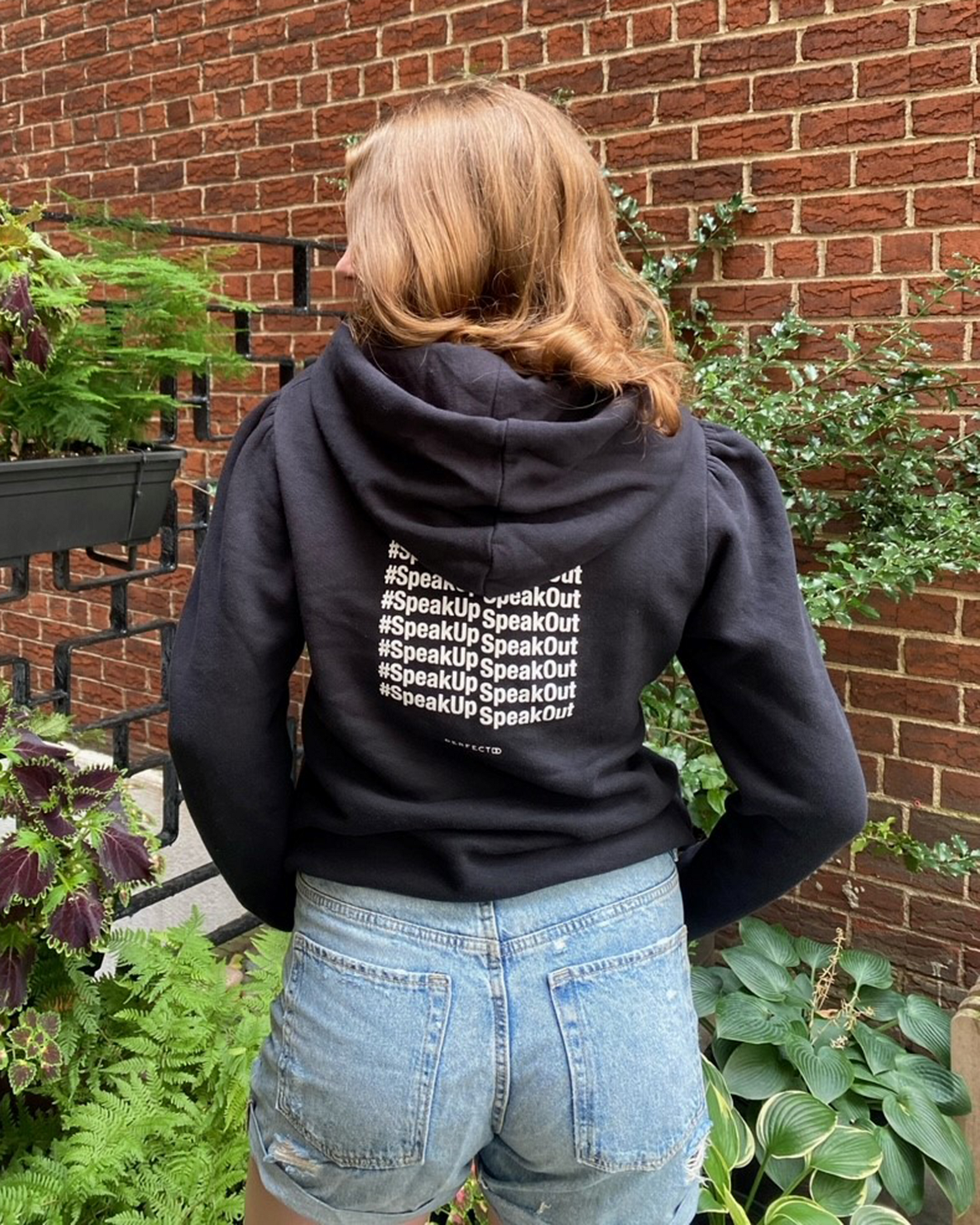 Back detail of black cotton fleece puff sleeve sweatshirt on model wearing blue jean shorts in front of brick wall and plants