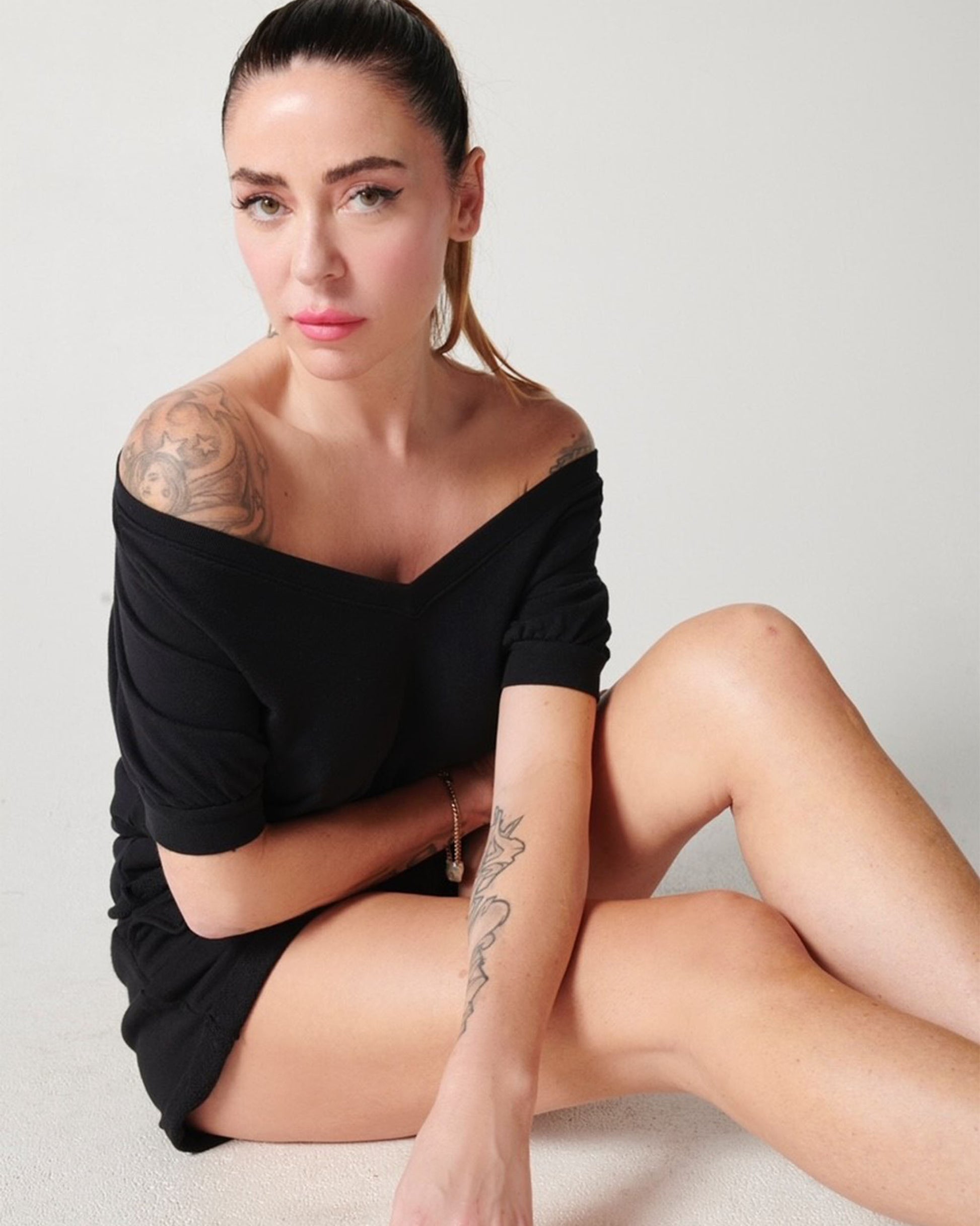 Black organic cotton v-neck sweatshirt and matching black pleated cotton sweat shorts on white model wearing the style off-the-shoulder while sitting on floor with arms crossed