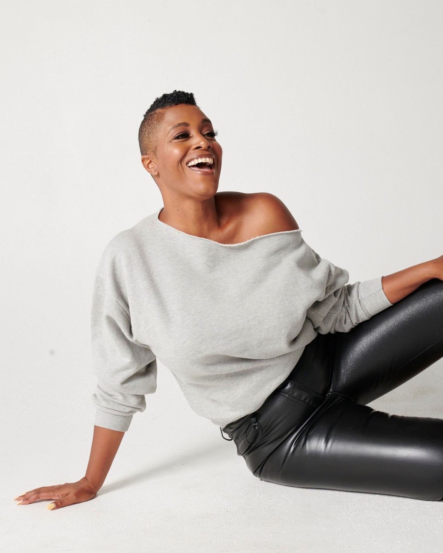 Heather grey cotton fleece off-the-shoulder sweatshirt on smiling model sitting down in black leather pants