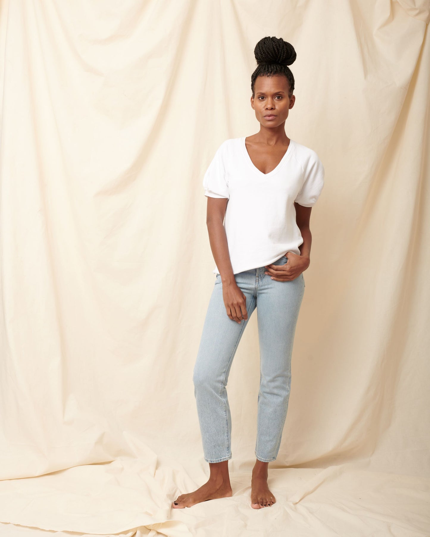Black model wearing white organic cotton v-neck sweatshirt and blue jeans with hand in pocket and bun in hair