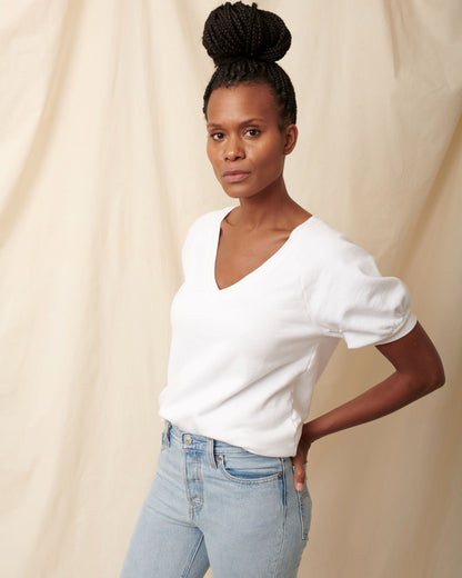 Black model wearing white organic cotton v-neck sweatshirt and blue jeans with hands in back pockets and bun in hair