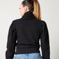 Back detail of black organic cotton turtleneck on model in blue jeans and ponytail sitting on stool