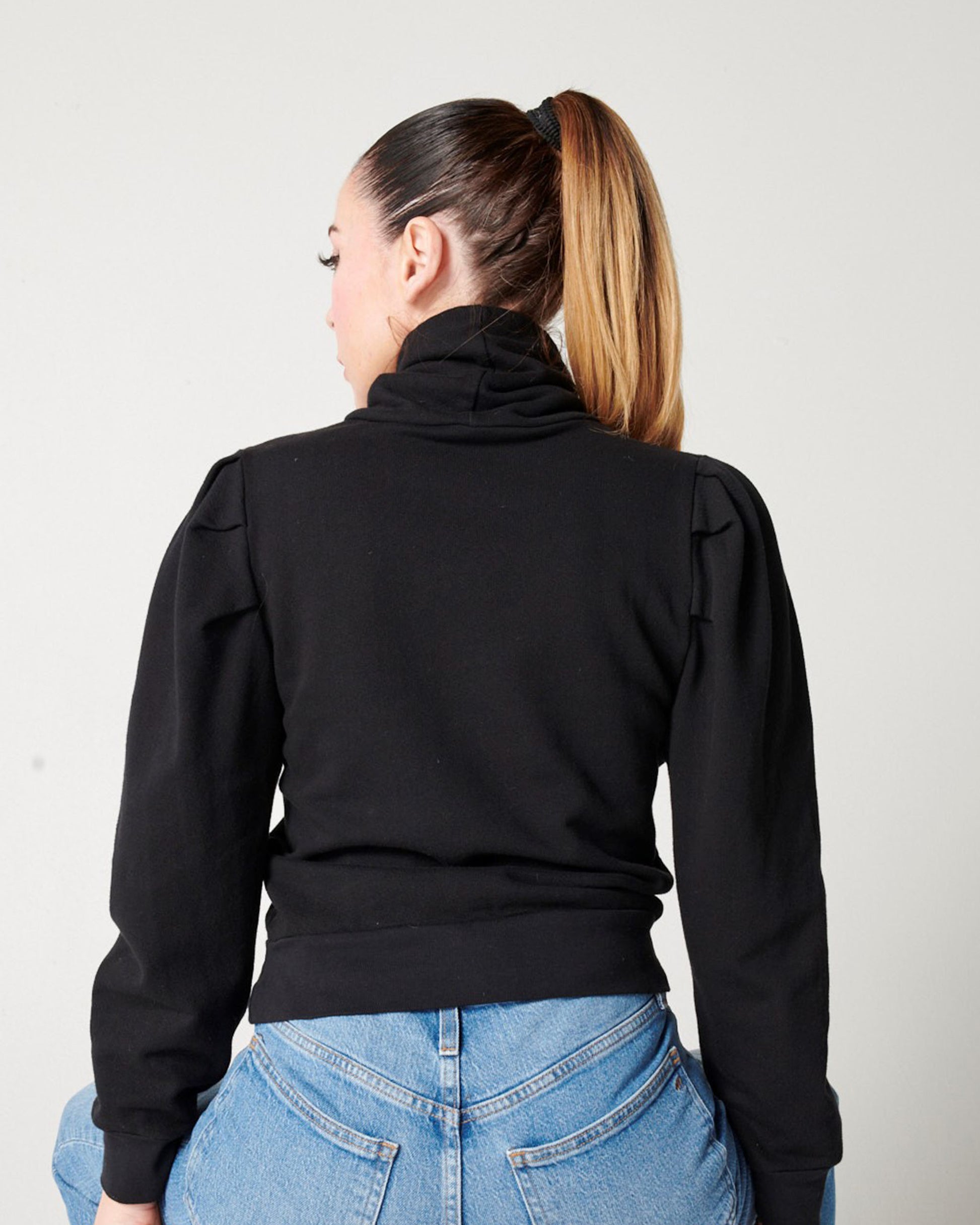 Back detail of black organic cotton turtleneck on model in blue jeans and ponytail sitting on stool
