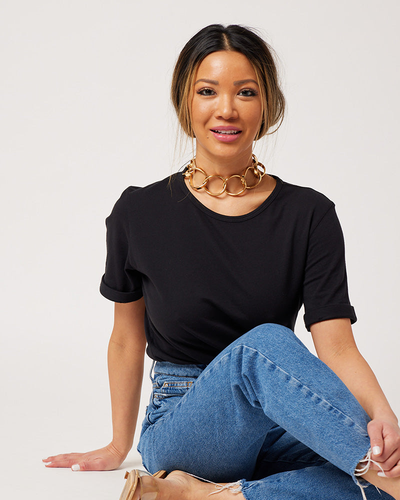 Black cotton rolled sleeve tshirt on model sitting on floor wearing chunky gold necklace, blue jeans and clear sandals