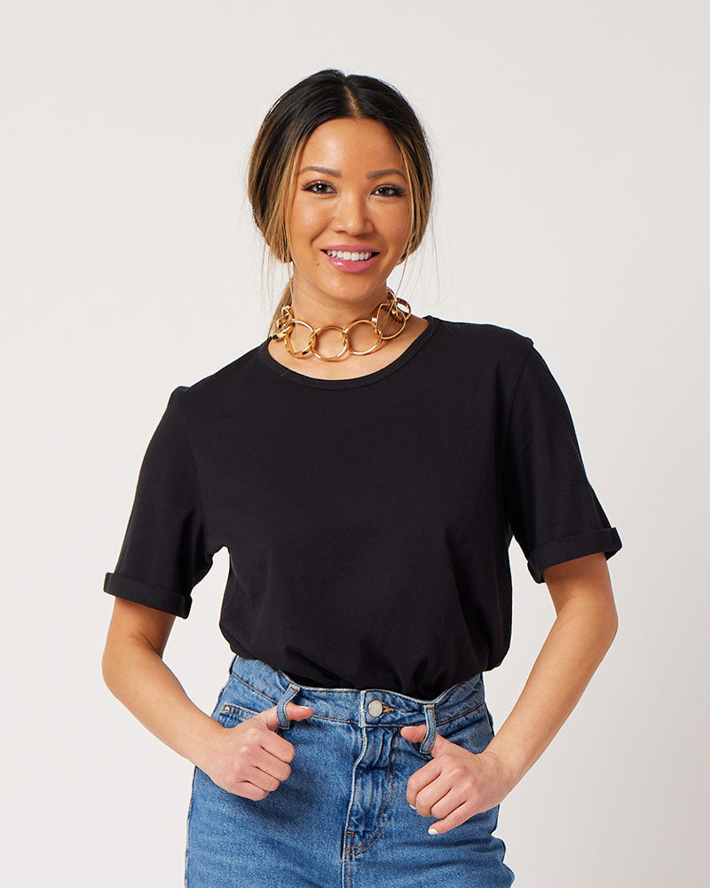 Black cotton rolled sleeve tshirt on smiling asian model wearing chunky gold necklace, blue jeans and clear sandals with hands in belt loops