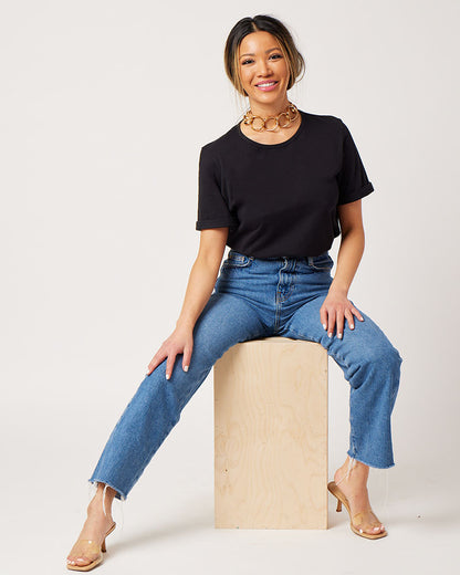 Black cotton rolled sleeve tshirt on smiling asian model sitting on stool wearing chunky gold necklace, blue jeans and clear sandals