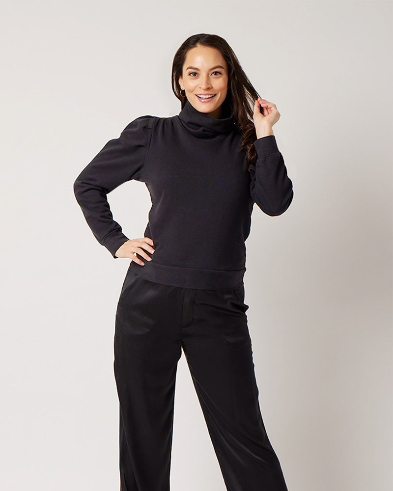 Black organic cotton turtleneck on smiling pregnant model in black silk pants with hand on hip and other holding hair