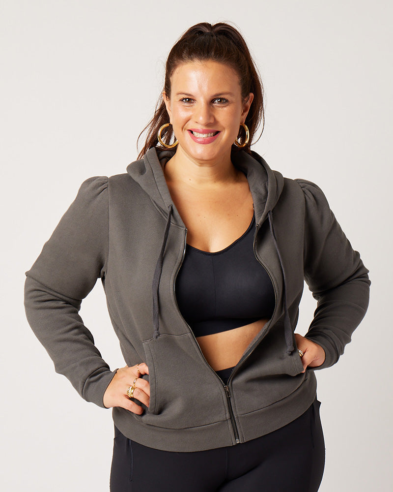 Grey cotton fleece puff sleeve zip up sweatshirt partially zippeed on model wearing black recycled polyester sports bra, black leggings, ponytail and gold hoops with hands on hips