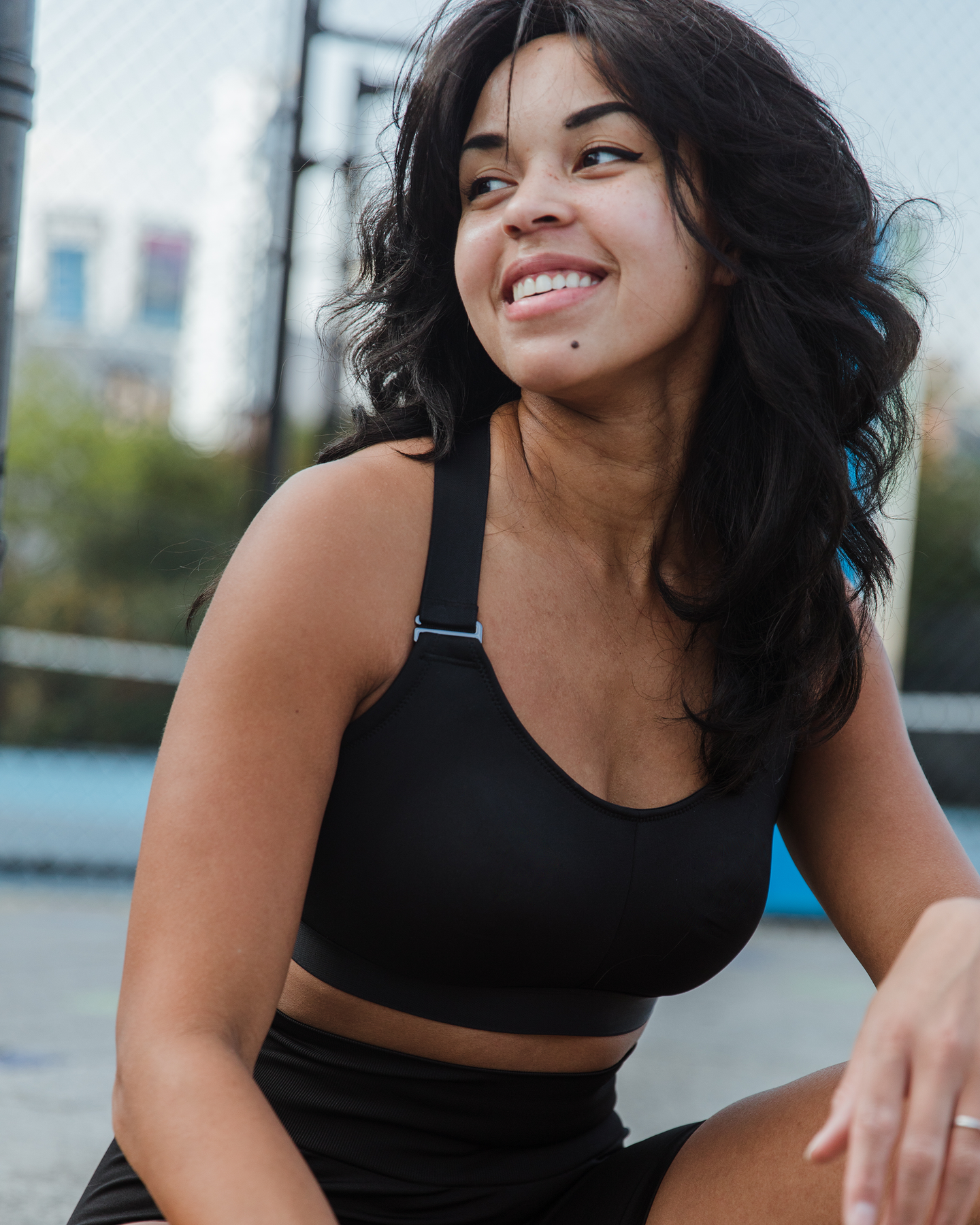 Black recycled polyester sports bra with adjustable shoulder strap on smiling fuller bust model sitting on tennis court, looking to side 