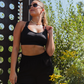 Black recycled polyester sports bra and black cotton pleated sweat shorts on white model in ponytail, gold jewelry and sunglasses outside holding onto shoulder adjustable strap