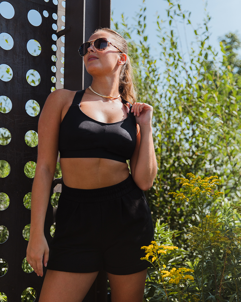 Black recycled polyester sports bra and black cotton pleated sweat shorts on white model in ponytail, gold jewelry and sunglasses outside holding onto shoulder adjustable strap