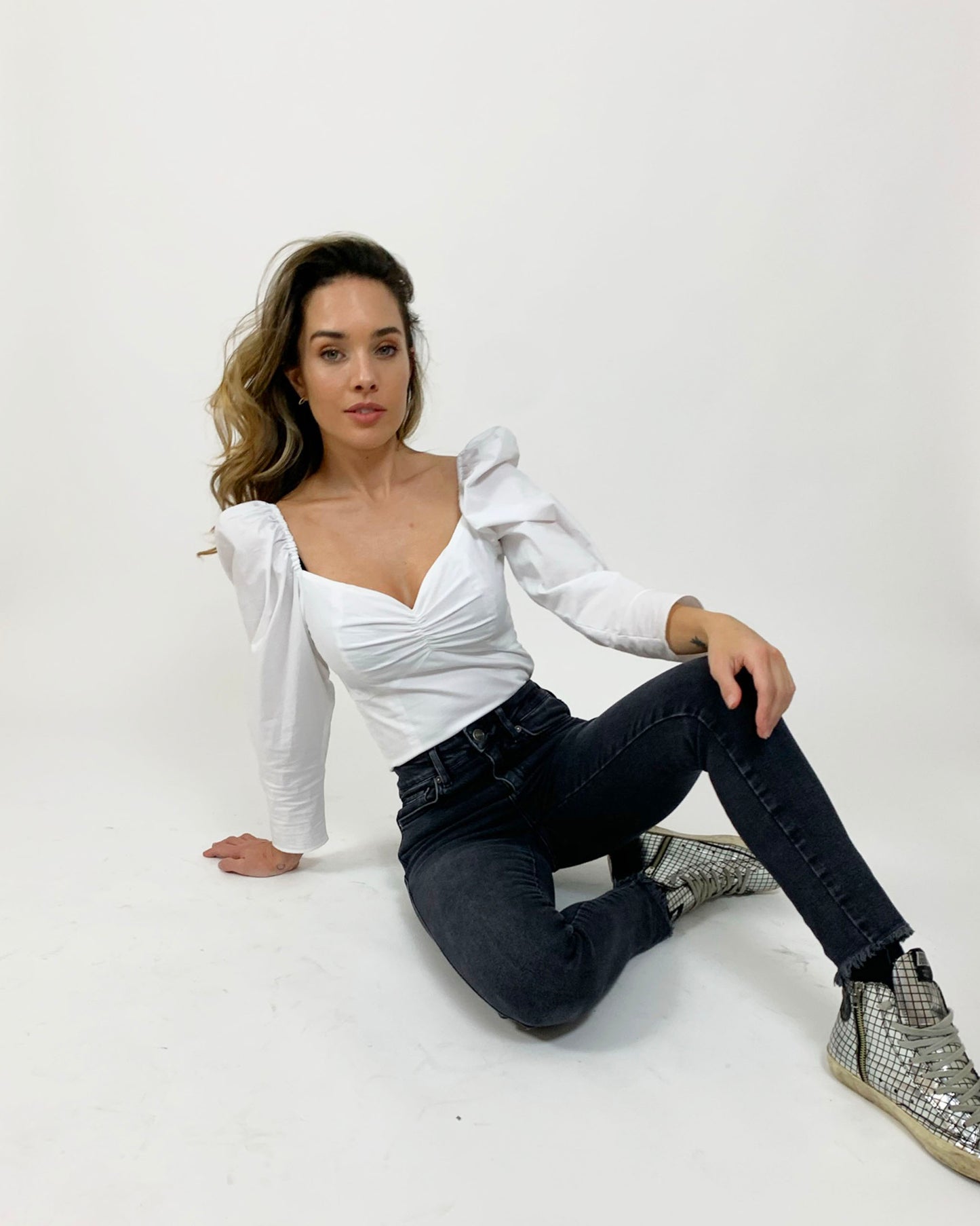 White cotton top with sweetheart neckline on brunette model sitting on floor in dark jeans and silver golden goose sneakers