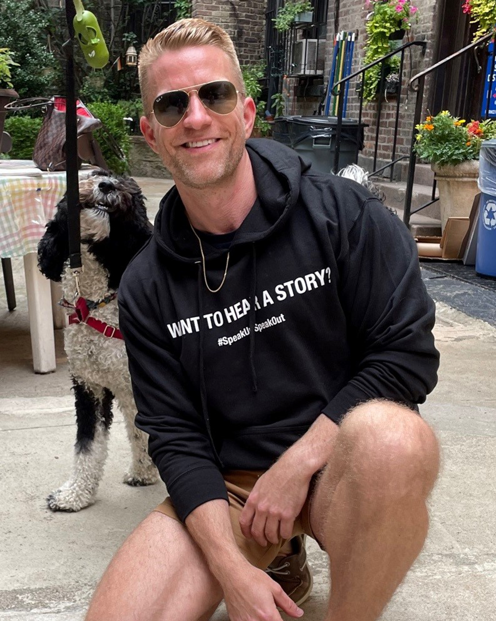 Black unisex long sleeve sweatshirt on blonde male model wearing sunglasses kneeling down next to black and white dog