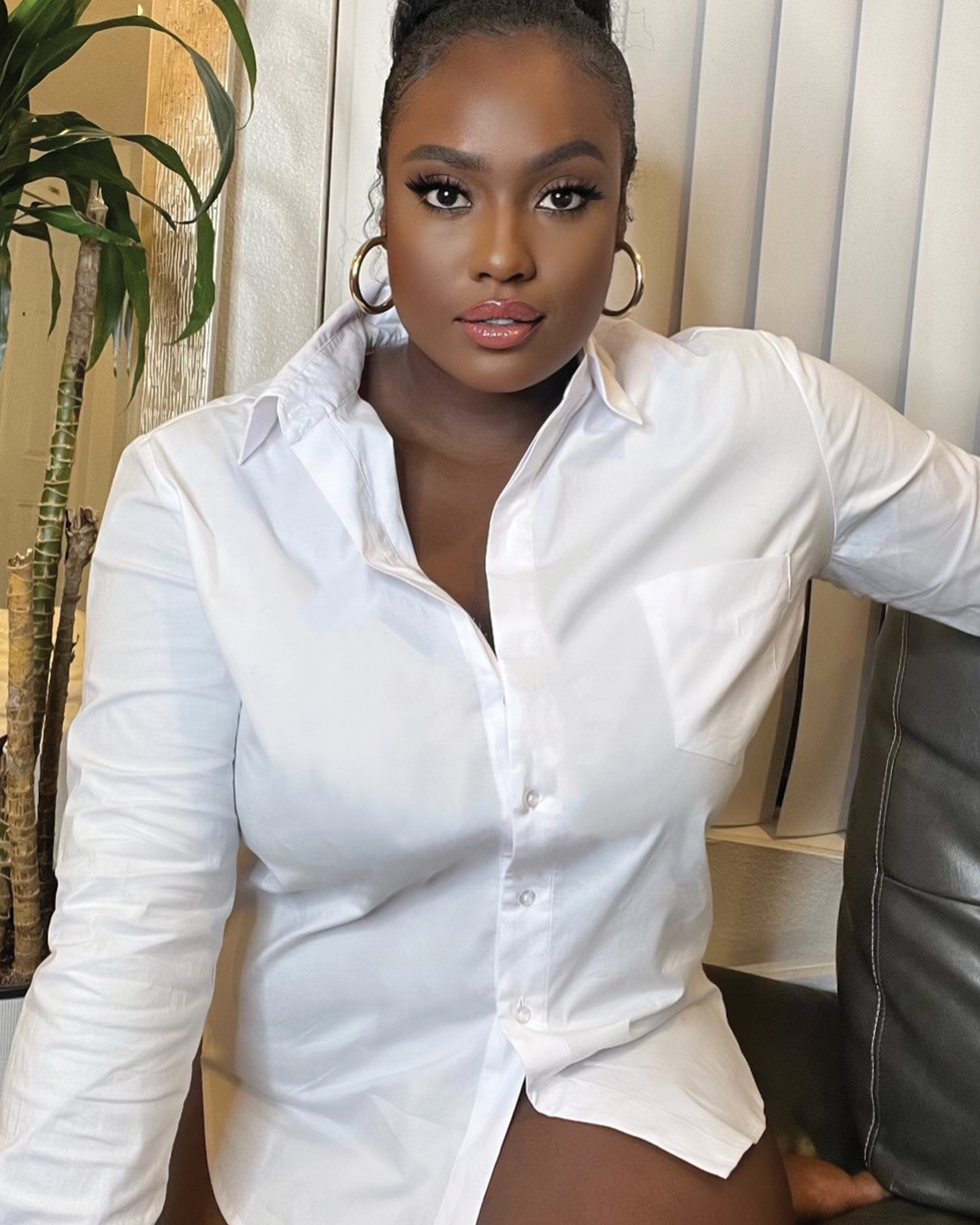 White organic cotton long sleeve button down on black model in bun and hoop earrings sitting on leather couch