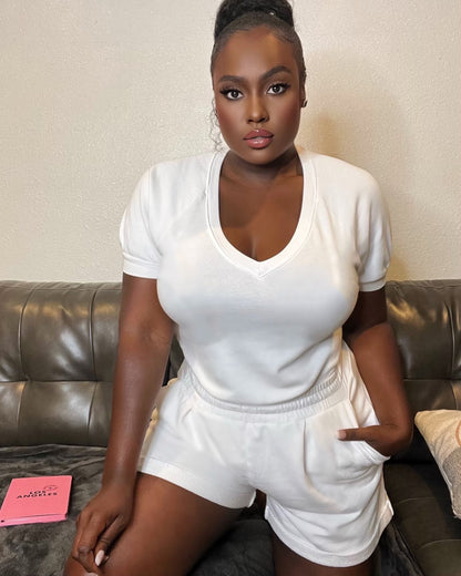 Black model wearing white organic cotton v-neck sweatshirt with matching white cotton pleated sweat shorts while kneeling on leather couch